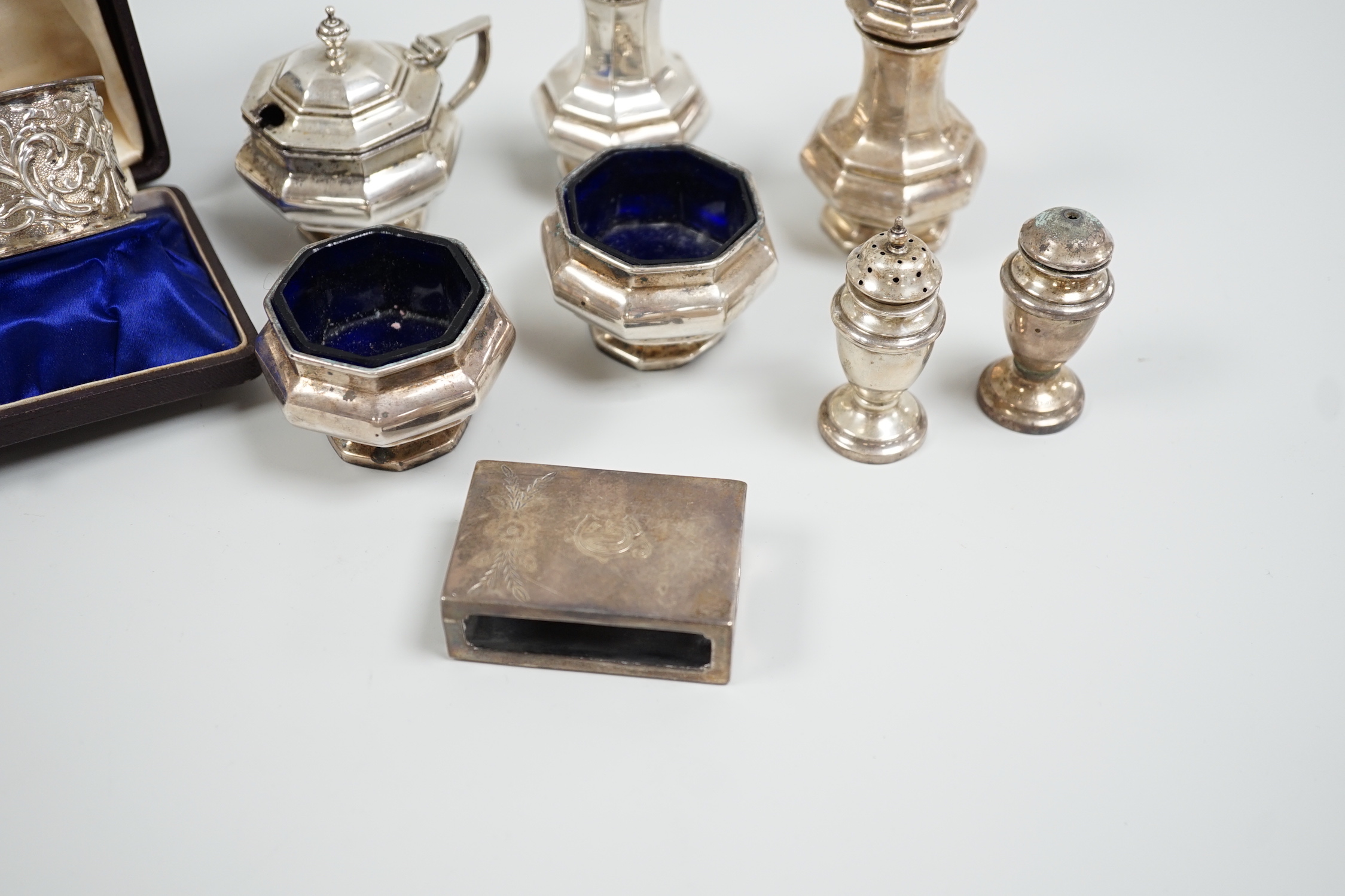 A cased pair of Edwardian embossed silver napkin rings, Birmingham, 1904, a five piece silver condiment set, Birmingham, 1926, two plated condiments and a match sleeve.